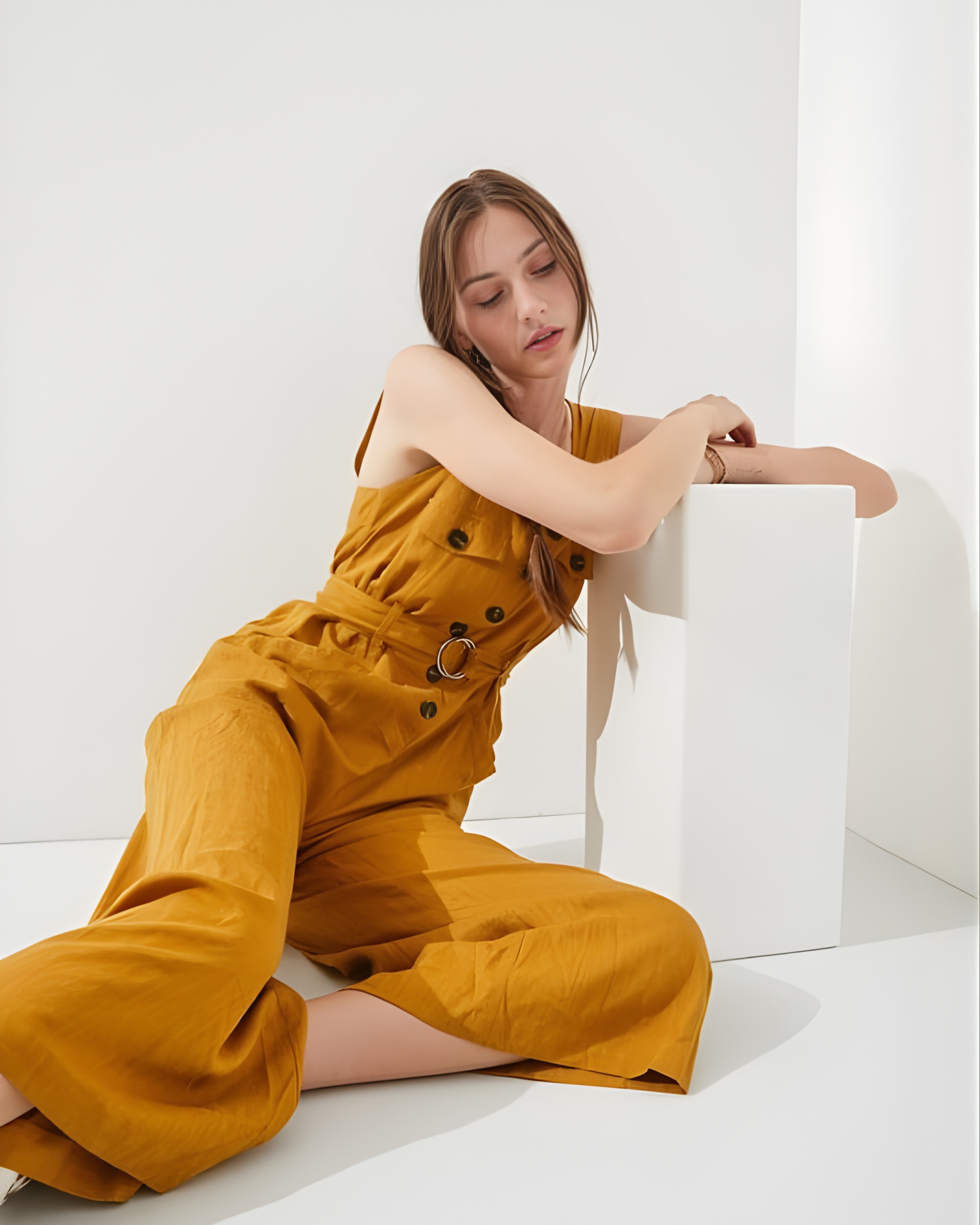 Woman in mustard yellow jumpsuit sitting on a white surface with a white background